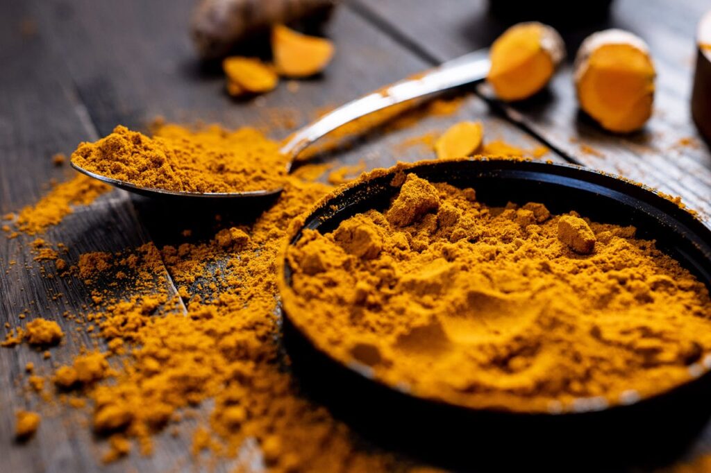 Pure Turmeric Powder | High Curcumin Haldi | TAVRAJ Close-up of vibrant turmeric powder with root on rustic wooden table, emphasizing texture and color.